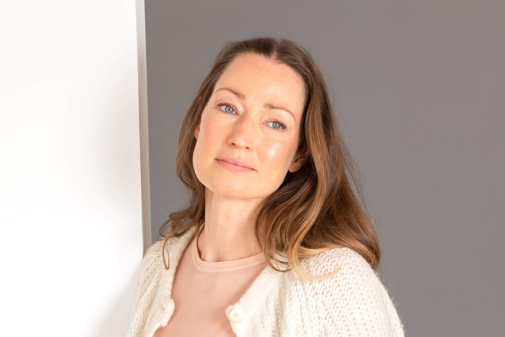 Frau, Alter Mitte 40, dunkel-blonde lange Haare, Blick frontal,Gesicht und Oberkörper frontal zu sehen, leichtes Lächeln, blaue Augen, steht, graue Wand im Hintergrund, weiße Strickjacke offen, rosé färbendes Oberteil, Nahaufnahme