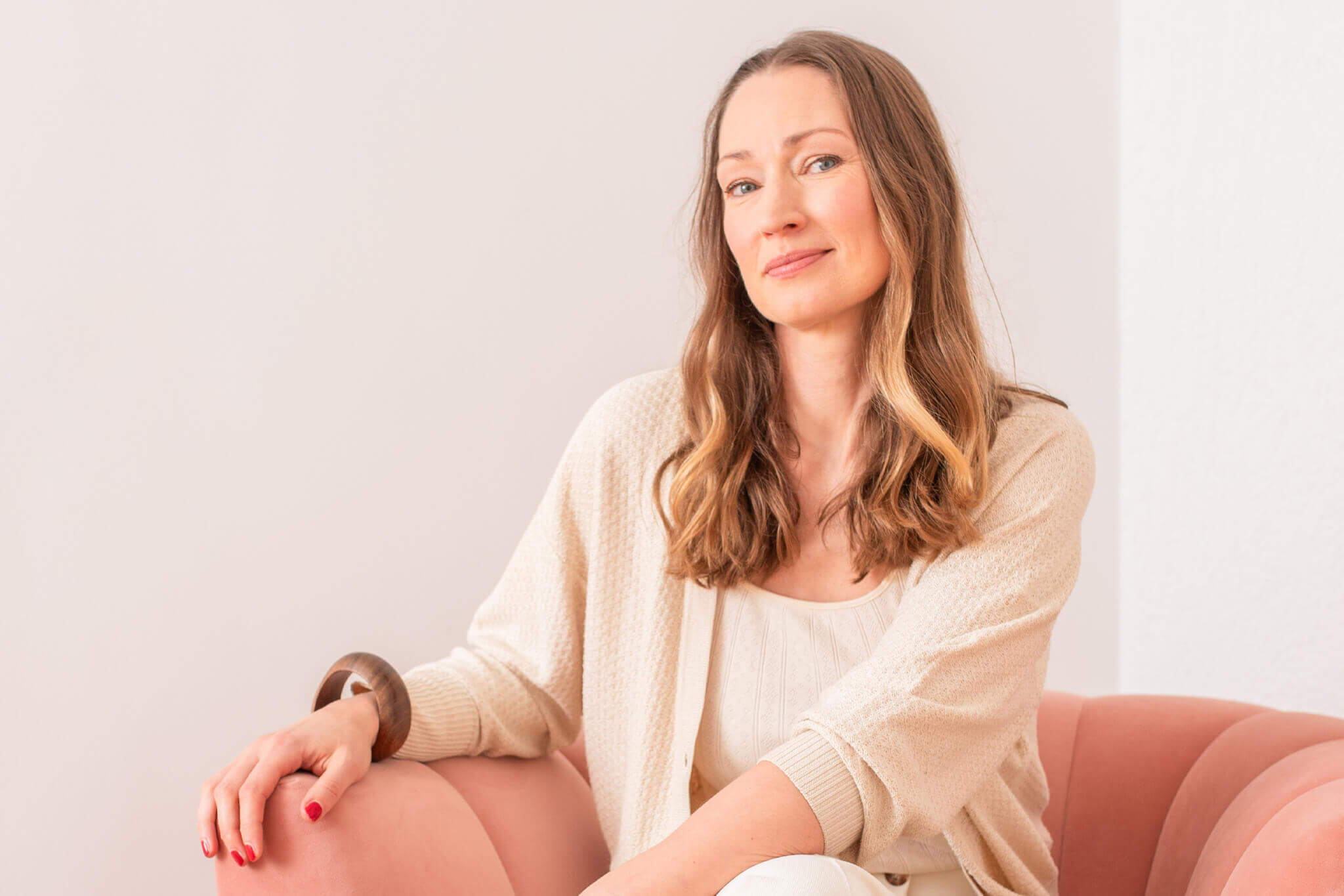 Frau, Alter Mitte 40, dunkel-blonde lange Haare, Blick direkt in die Kamara, Gesicht und Oberkörper frontal zu sehen, leichtes Lächeln, blaue Augen, sitzt auf rosa färbenden Sessel, helle Wand im Hintergrund, naturweißes Top mit naturweißes dünnen Strickjacke, linker Arm (vom Betrachter aus) liegt auf Sessel auf, brauner Holzarmreif, roter Nagellack, Nahaufnahme