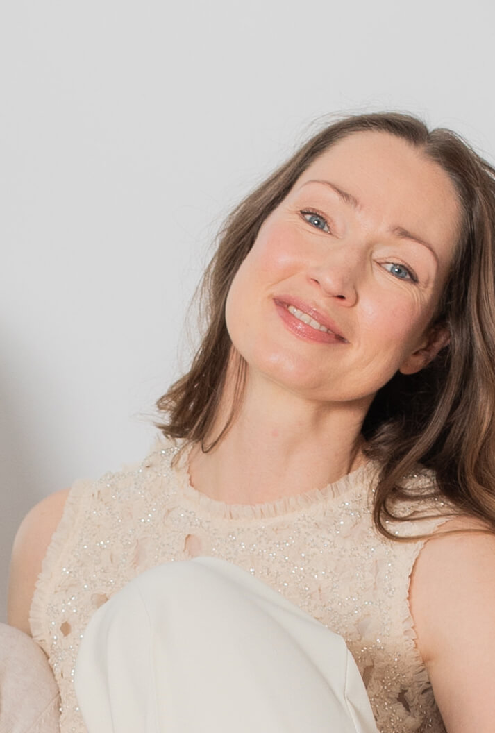 Frau, Alter Mitte 40, dunkel-blonde lange Haare, Gesicht frontal zu sehen, leichtes Lächeln, blaue Augen, helles Oberteil, leicht glitzernd, Schultern und Oberarme unbedeckt, weiße Stoffhose, Ausschnitt Gesicht und Teile des Oberkörpers und die angewinkelten Beine sind zu sehen, hell-graue Wand im Hintergrund